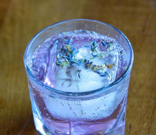 A clear glass filled with a purple drink, ice cubes, and garnished with dried flowers, resting on a wooden surface.
