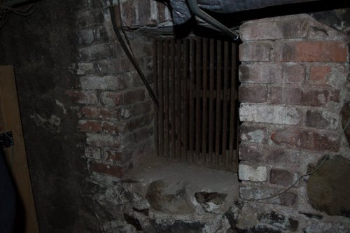 A dimly lit brick wall with a barred window, revealing a dusty, shadowy space behind it.