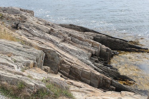 Rocky shoreline with layered stone formations and seaweed, overlooking calm waters.