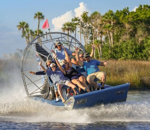A group of excited people enjoying an airboat ride, splashing through water with lush greenery in the background.