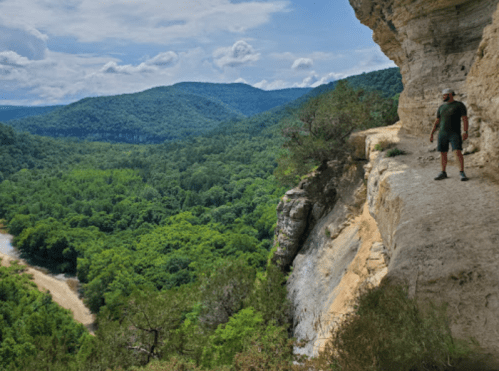 A person stands on a rocky ledge overlooking a lush green valley and river, surrounded by mountains under a blue sky.