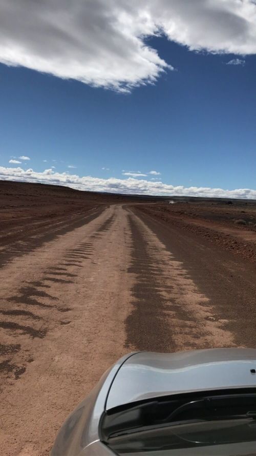 A dirt road stretches into the distance under a blue sky with scattered clouds. A car's hood is visible in the foreground.
