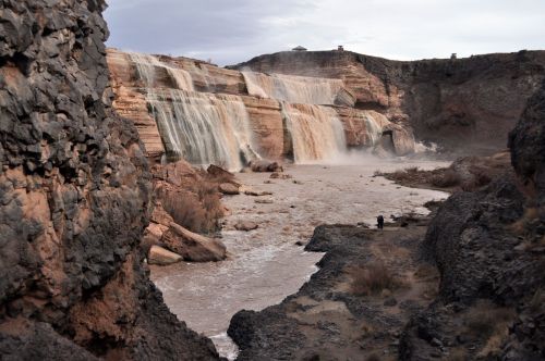 A stunning waterfall cascades over rocky cliffs into a river, surrounded by rugged terrain and a cloudy sky.