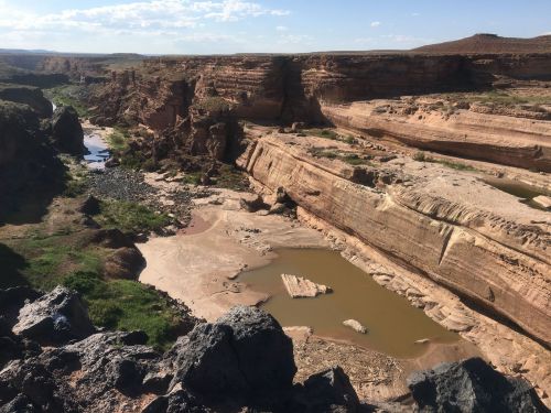 A scenic view of a canyon with rocky cliffs, a winding river, and patches of green vegetation under a blue sky.