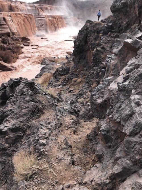 A person walking along rocky terrain near a river with flowing water in a natural landscape.