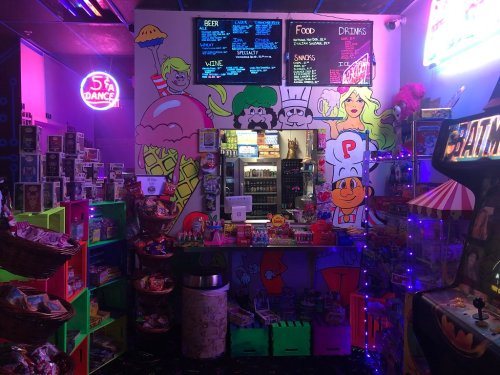 Colorful arcade snack bar with neon signs, cartoon murals, and shelves filled with candy and snacks.