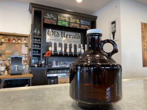 A brown glass growler sits on a counter in a brewery, with a bar and shelves in the background.