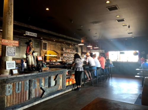 A rustic bar interior with patrons seated at the counter, a cowboy statue, and a variety of drinks displayed.