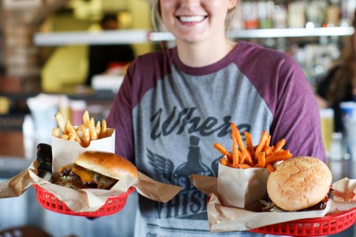 A smiling woman holds two baskets of burgers and fries in a casual dining setting.