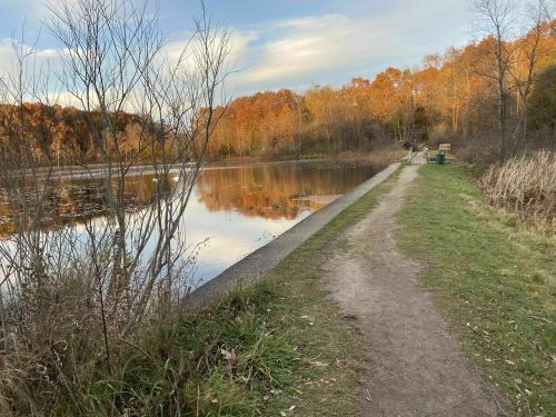 A serene path along a calm lake, surrounded by autumn foliage and trees, under a clear sky.