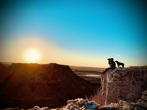 A person and a dog sit on a cliff, watching a sunset over a vast landscape.