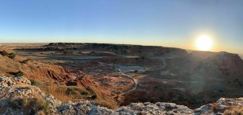 A panoramic view of a rugged landscape at sunset, featuring red earth and distant hills under a clear sky.