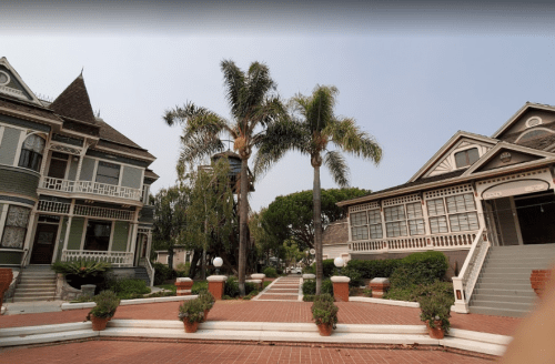 A charming street scene featuring Victorian-style houses and palm trees, with a brick pathway and landscaped gardens.