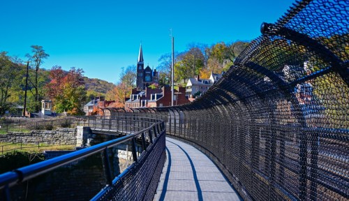 A winding bridge with a mesh railing, overlooking a town with colorful trees and a church steeple in the background.