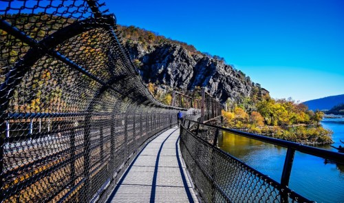 A winding pedestrian bridge with a mesh fence, surrounded by autumn foliage and rocky hills under a clear blue sky.