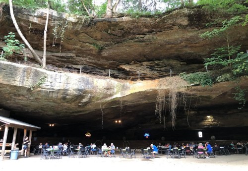 A cave-like setting with diners seated at tables, surrounded by rock formations and greenery above.