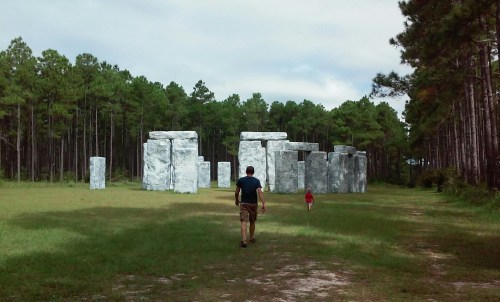 A man and a child walk towards a replica of Stonehenge in a grassy area surrounded by tall trees.