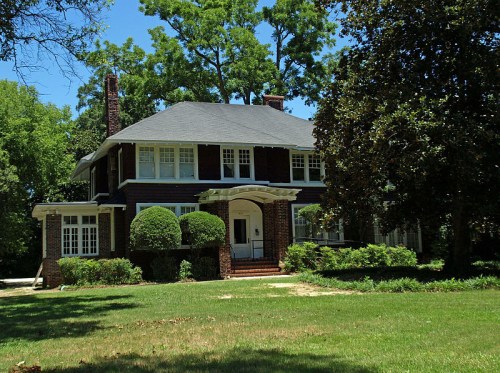 A large, two-story brick house with a gray roof, surrounded by lush greenery and trees on a sunny day.