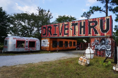 A colorful drive-thru entrance with a vintage trailer and graffiti art, surrounded by trees and open space.