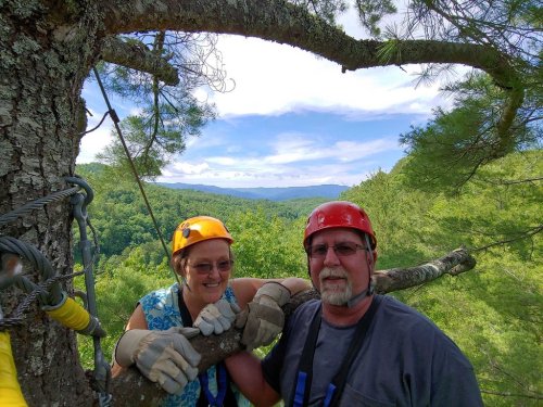 Two people in helmets smile while holding onto a tree branch, surrounded by lush green mountains and a blue sky.