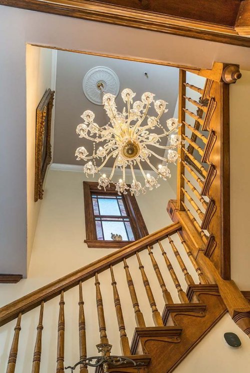 A view of a wooden staircase leading up to a chandelier and a window, showcasing elegant interior design.