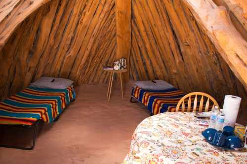 Interior of a cozy wooden hut with two beds, a small table, and colorful blankets. Natural light filters in.