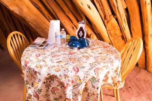 A wooden table with a floral tablecloth, two chairs, water bottles, and blue dishes against a rustic wooden wall.