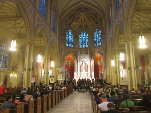 Interior of a grand church with stained glass windows, filled with people seated and a choir performing at the front.