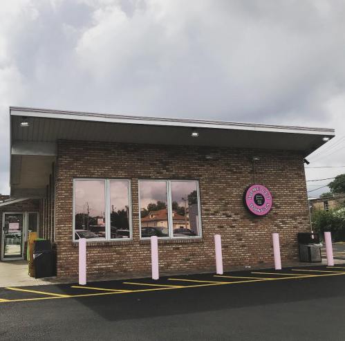 A brick building with large windows and a pink donut sign, set against a cloudy sky. Parking lot in front.