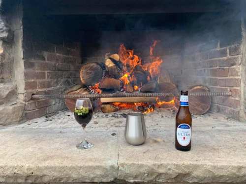 A cozy fireplace with a roaring fire, featuring a glass of dark beer and a bottle of light beer on a stone surface.