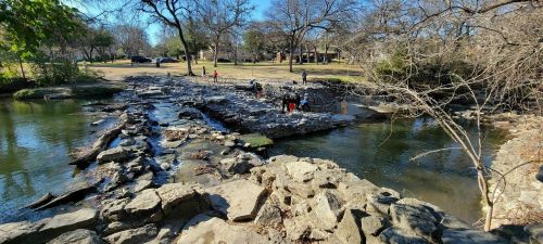 A rocky riverbank with a stone structure, surrounded by trees and people exploring the area on a sunny day.