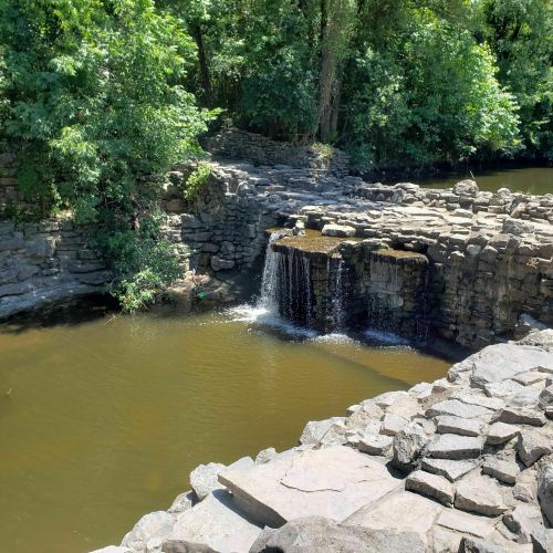 A serene waterfall cascades over stone formations into a calm, greenish-brown pool surrounded by lush trees.