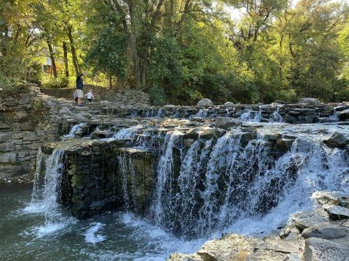 A serene waterfall cascades over rocky ledges, surrounded by lush greenery and a person standing nearby.