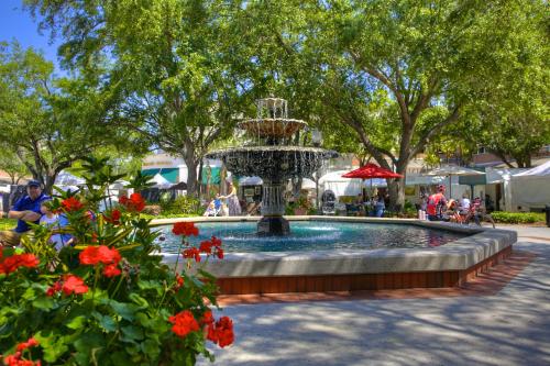 A decorative fountain surrounded by greenery and flowers, with people enjoying a market in the background.