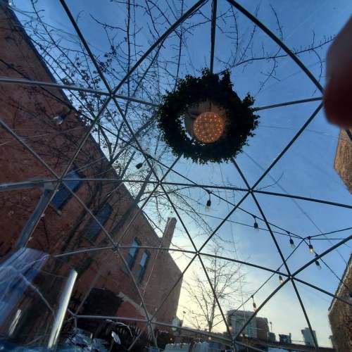 View from inside a transparent dome, featuring a circular light fixture and greenery, with a blue sky above.