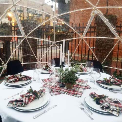 A festive dining setup inside a transparent dome, featuring a table with plaid decor and elegant tableware.