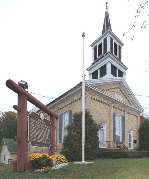 Historic church building with a tall steeple, surrounded by trees and flowers, next to a historical marker.