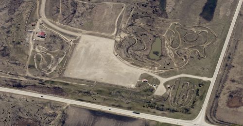 Aerial view of a dirt track with winding paths, a pond, and surrounding fields, near a road.