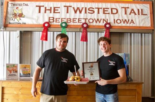 Two men in black shirts hold an award and a plate of food in front of a banner for The Twisted Tail in Bebeeetown, Iowa.