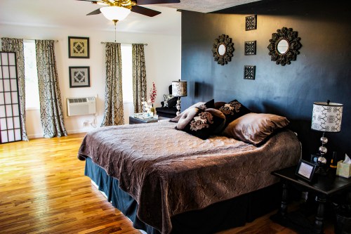 Cozy bedroom with a large bed, decorative pillows, dark walls, and elegant curtains, featuring wooden floors and stylish lamps.
