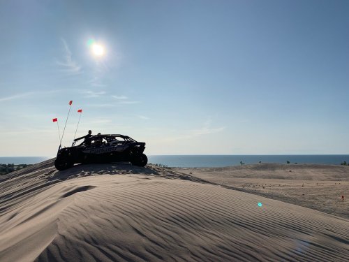 A dune buggy parked on a sandy hill with the ocean in the background and a clear blue sky above.