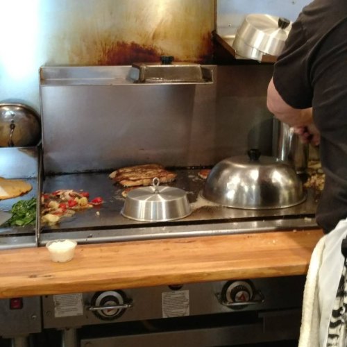 A chef cooking on a griddle with vegetables and a covered dish, in a restaurant kitchen setting.