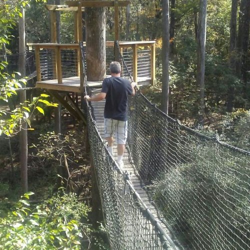 A person walks on a suspended bridge leading to a treehouse in a wooded area.