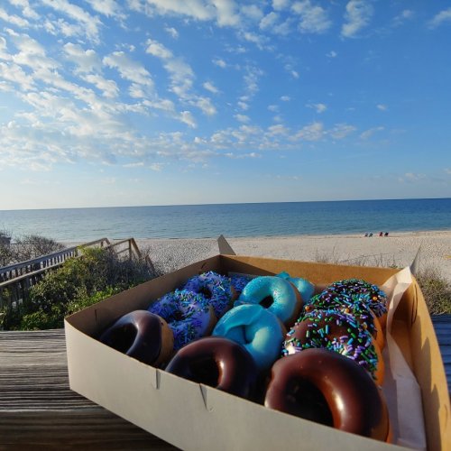 A box of colorful donuts sits on a wooden railing with a beach and blue sky in the background.