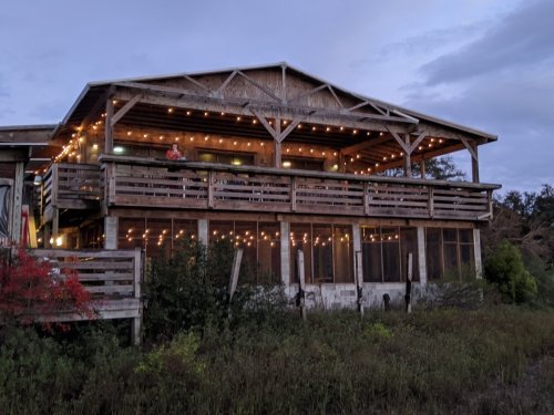 A rustic wooden house with a large balcony, adorned with string lights, surrounded by greenery at dusk.