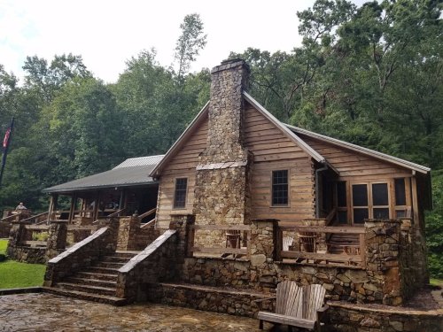 A rustic wooden cabin with a stone chimney, surrounded by trees and a stone patio with seating.