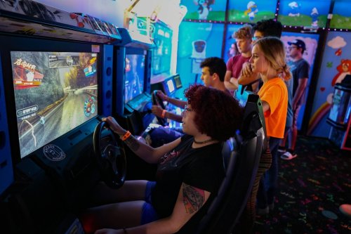 A group of friends plays racing arcade games in a colorful game room, with one person driving and others watching.