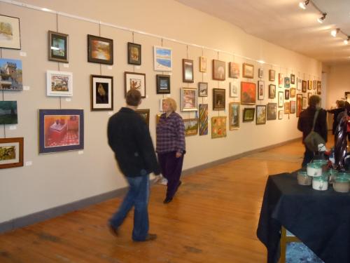 Two people walk through an art gallery, admiring various framed artworks displayed on the walls.