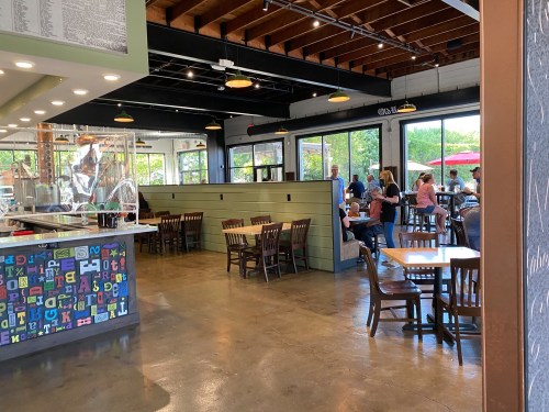 Interior of a casual restaurant with wooden tables, chairs, and large windows, bustling with customers.