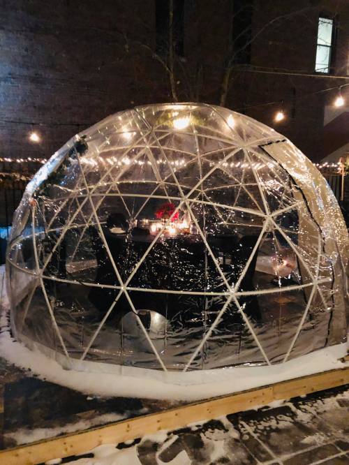 A clear geodesic dome with a wooden base, illuminated by string lights, set in a snowy outdoor area.